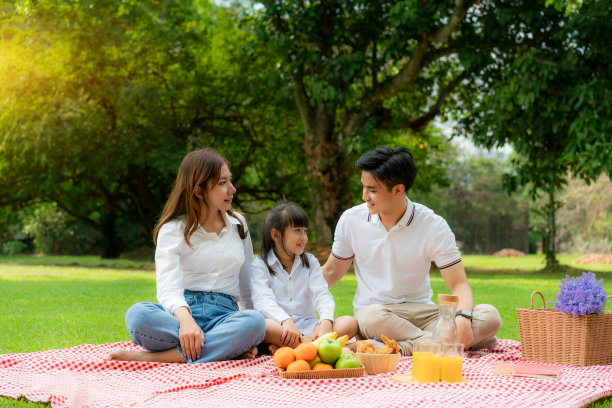 亚洲青少年家庭愉快的假日野餐时刻在公园与母亲和女儿看着父亲和微笑愉快度过假期时间在绿色花园与水果和食物。图片下载