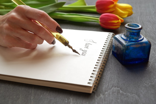 手持旧钢笔在笔记本上写“我爱你”，黑色背景，鲜花和墨水瓶图片下载