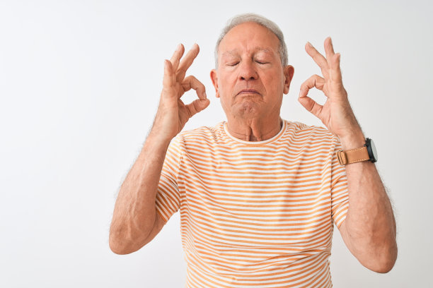 白发老人身穿条纹t恤，站在孤立的白色背景下，闭着眼睛微笑着做冥想手势。瑜伽的概念。图片下载