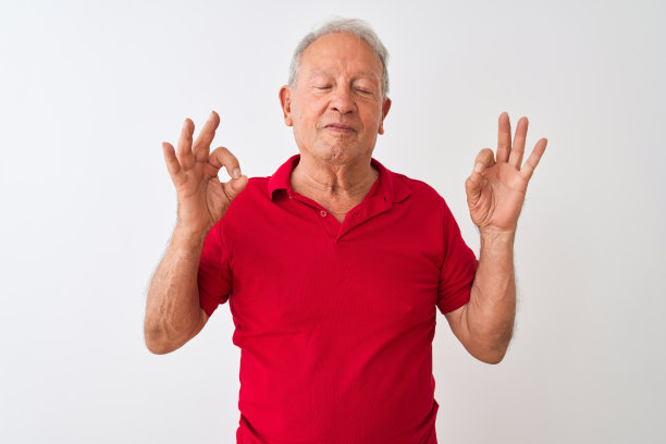 年长的灰头发男子穿着红色马球站在孤立的白色背景放松和微笑，闭上眼睛做冥想手势与手指。瑜伽的概念。图片下载