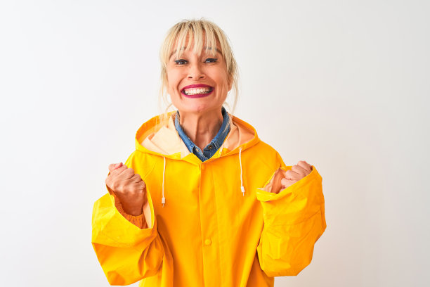 身着黄色雨衣的中年妇女站在孤立的白色背景下，举起双臂，睁大眼睛庆祝成功。赢家的概念。图片下载