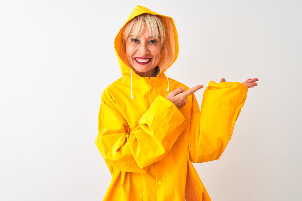 一位中年妇女穿着雨衣，带着兜帽，站在孤立的白色背景下，对着镜头惊讶地微笑着，同时用手指着镜头。图片下载