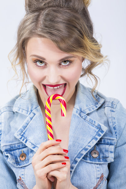 美食创意概念：吃着大红色黄色拐杖糖的惊讶白人金发女孩的肖像（竖版照片）图片下载