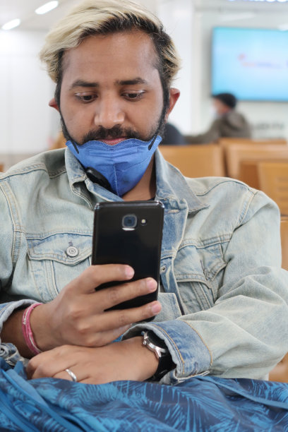 这是一幅印度亚裔男子坐在候机室里玩手机的照片，他戴着口罩来保护冠状病毒、污染、全球卫生紧急情况下的口罩、呼吸问题、病毒性肺炎、SARS、疾病、检疫和疾病图片下载