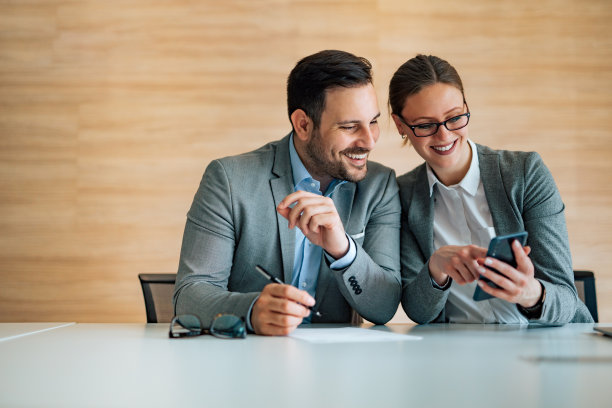 两个微笑的商务人士坐在办公桌旁一起看智能手机图片下载