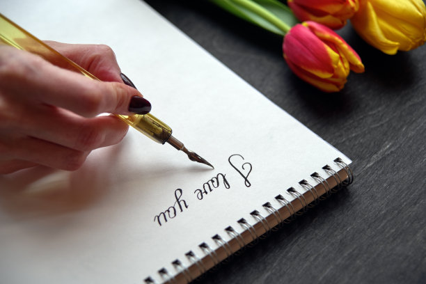 手持旧钢笔在笔记本上写“我爱你”，黑色背景，鲜花和墨水瓶图片下载