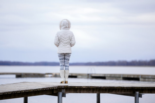 年轻女子穿着白色温暖的衣服和靴子，独自站在木桥上，凝视着湖面。连帽的女孩。宁静的自然氛围。在冬天享受新鲜空气。后视图。图片下载