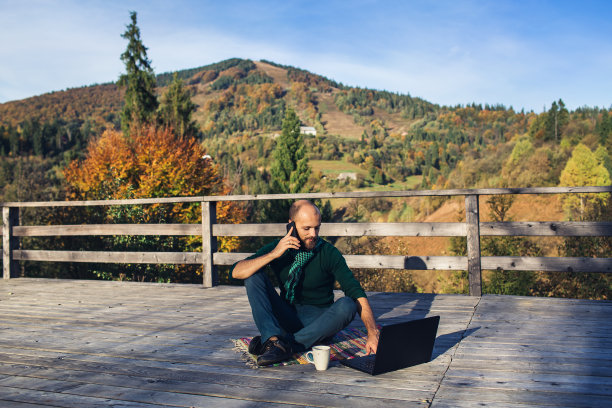 络腮胡男子坐在山上的露台上，用笔记本电脑打电话图片下载