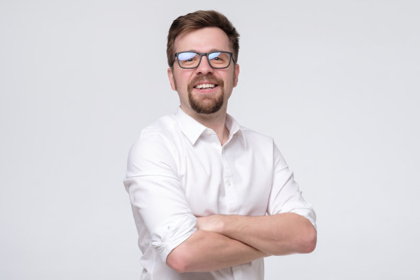 戴着眼镜的年轻自信的男人微笑着站在白色的墙上。图片下载