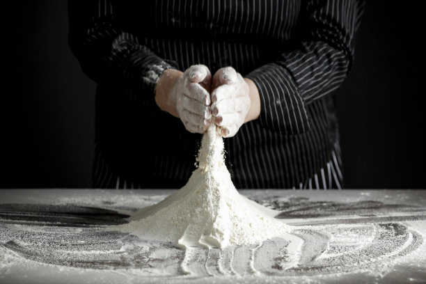 在晨曦中，厨房的桌子上放着面粉，背景是黑色的。女人们用手筛面粉，做面团。图片下载