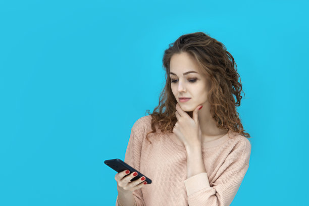 一个年轻美丽的女人的肖像穿着运动衫拿着一个手机在她的手，看着它深思熟虑在蓝色的背景图片下载