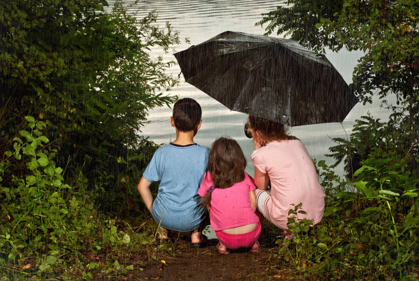 孩子们坐在河边的雨中，女孩和男孩在一把黑伞下，孩子们在湖边的雨中淋湿。图片下载