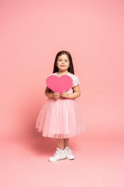 穿着薄纱裙，拿着红色纸质爱心，对着镜头微笑的时尚小女孩，粉色背景图片下载