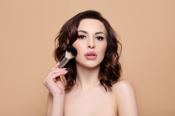 年轻漂亮的微笑女人在米色背景上拿着化妆刷，美容院概念，清新干净的皮肤图片下载