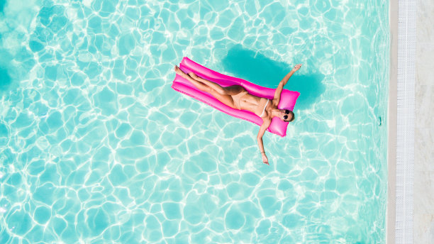 在泳池的漂浮物上休息和日光浴的迷人女性，俯视航拍照片。比基尼泳装的年轻女人漂浮在粉色充气床垫上图片下载
