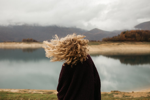 快乐的女孩，一头金色的卷发独自在湖面上跳舞，她的头发因为风的流动而飞舞，像鸟儿一样自由。照片中的女孩与卷发站在后面的相机在湖的背景。图片下载