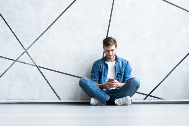 一个微笑的年轻男子坐在地板上，用智能手机对着灰色的墙壁。图片下载