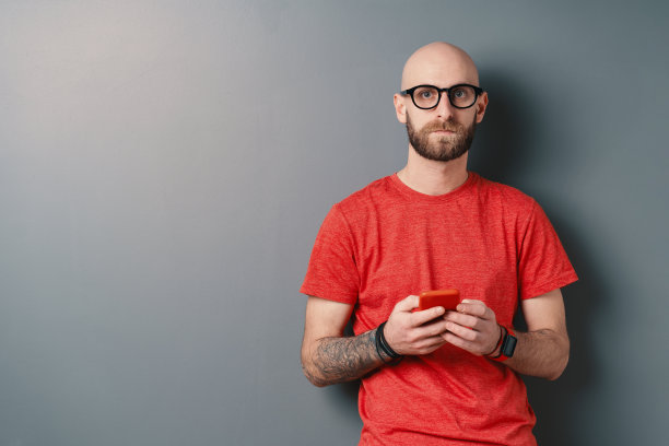 英俊的光头白种人胡子，眼镜，红色t恤拿着手机在灰色的演播室背景图片下载