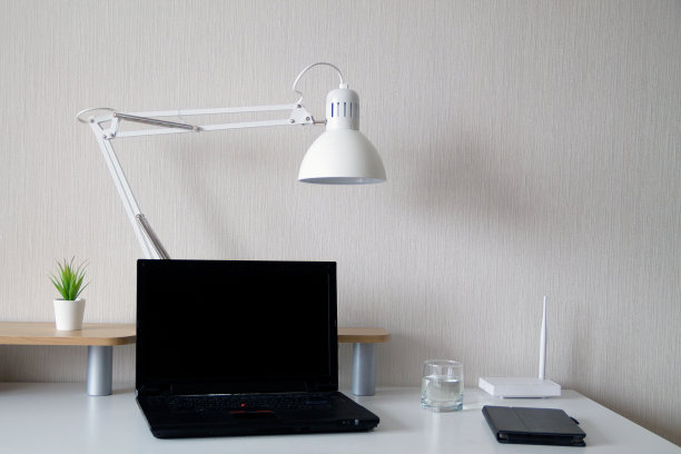 现代办公室内。白色的台灯和一株小植物，一杯水和黑色的笔记本电脑放在白色的桌子上。桌面的组织。素材图片
