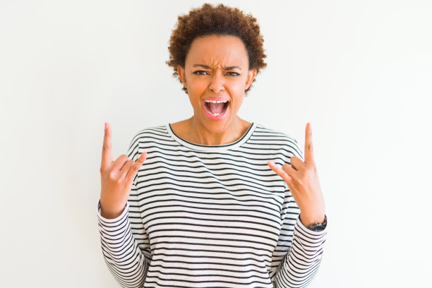 年轻漂亮的非裔美国妇女穿着条纹毛衣在白色背景下，以疯狂的表情喊叫，双手举起做摇滚标志。音乐明星。重金属概念。图片下载