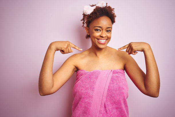 非洲裔女人，浴后裹着浴巾，在粉色背景前，面带微笑，自信地用手指指向自己，自豪而快乐。图片下载