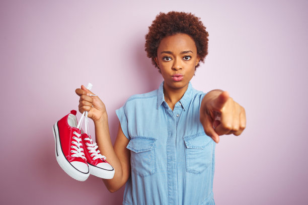 非洲裔女人在粉色背景前拿着红色休闲运动鞋，用手指着相机图片下载