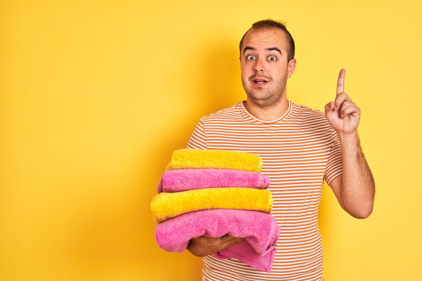 年轻的店主拿着折叠的毛巾站在孤立的黄色背景惊讶地提出一个想法或问题，用幸福的脸指着手指，第一个图片下载