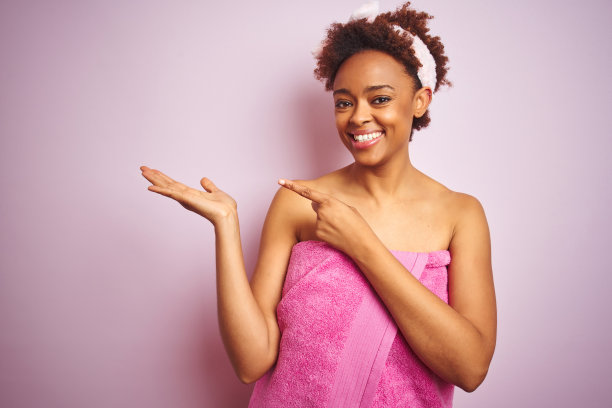 沐浴后裹着浴巾的非洲裔美国女人在粉色背景前惊讶地对着镜头微笑，用手展示并用手指指向图片下载