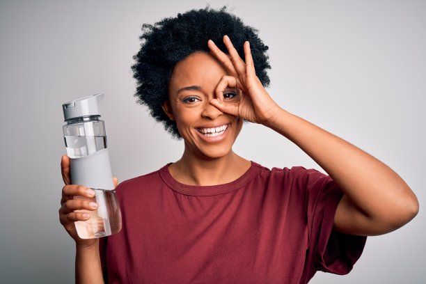 喝着瓶装水、面带微笑、用手在眼前做OK手势的年轻非裔美国蓬松发型女性图片下载