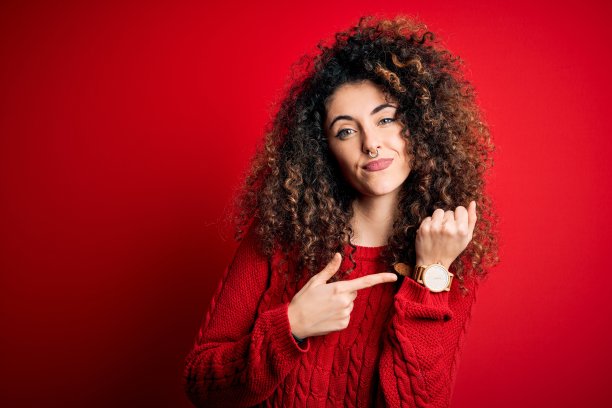 年轻漂亮的女子一头卷发，穿一件休闲的红毛衣，匆忙的指着看时间，不耐烦的看着镜头，用轻松的表情图片下载