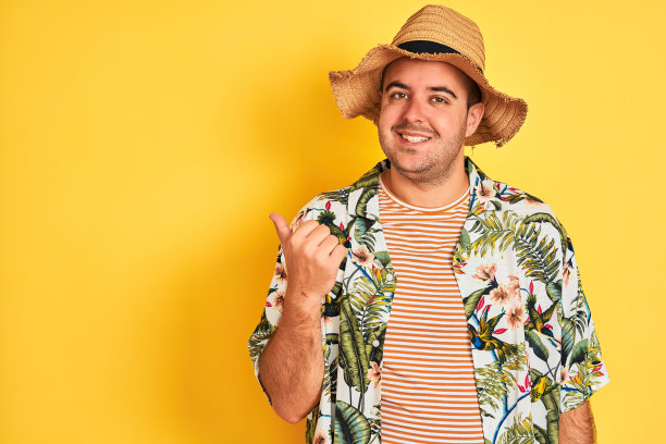 年轻男子度假穿夏季衬衫和帽子在孤立的黄色背景微笑与快乐的脸看和指着一边拇指向上。图片下载