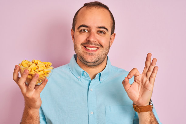 年轻男子拿着碗通心粉站在孤立的粉红色背景做ok手势的手指，优秀的象征图片下载