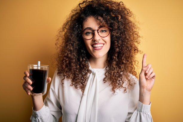 年轻漂亮的女人，一头卷发，戴着眼镜，喝着咖啡，惊讶于一个想法或问题，用快乐的脸指着手指，第一图片下载
