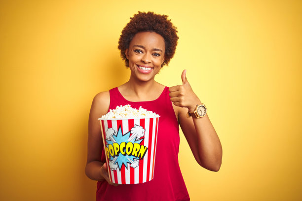 非洲裔美国妇女拿着一包爆米花在黄色孤立的背景下开心地笑着做ok的手势，用手指竖起大拇指，优秀的手势图片下载