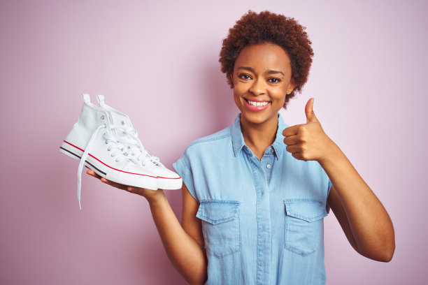 非裔美国女人拿着白色休闲运动鞋，在粉红色背景上高兴地笑着，做着OK的手势，竖起大拇指，表示很棒图片下载