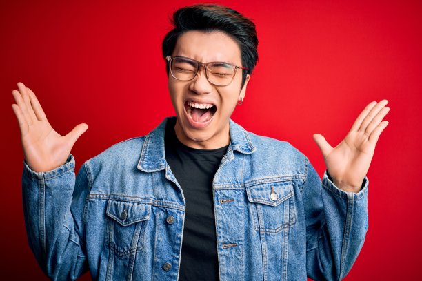 年轻帅气的中国男子身穿牛仔夹克，戴着眼镜，在红色背景下高举双臂，闭着眼睛兴奋地尖叫，为成功而疯狂。赢家的概念图片下载