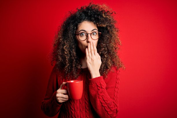年轻漂亮的女子，一头卷发，穿洞喝着红色的咖啡，捂着嘴用手捂着，因错误而震惊，表情恐惧，在沉默中害怕，秘密的概念图片下载