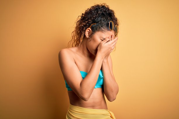 年轻美丽的美国女人度假穿着比基尼在孤立的黄色背景与悲伤的表情掩盖脸与手哭泣。抑郁症的概念。图片下载