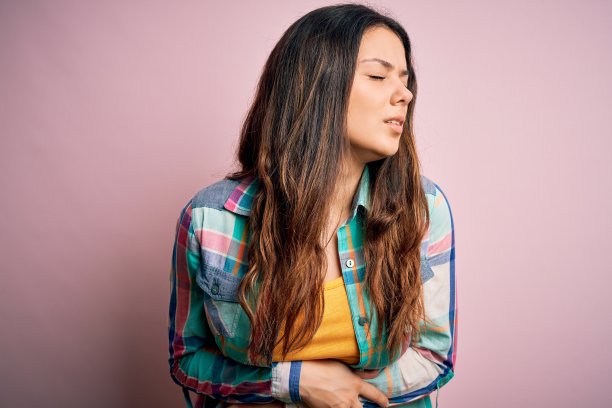 年轻美丽的黑发女子穿着休闲彩色衬衫站在粉红色的背景与手在胃部，因为恶心，痛苦的疾病感觉不舒服。疼痛的概念。图片下载