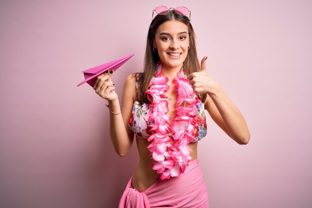 年轻美丽的女人在度假穿着比基尼和夏威夷花环拿纸飞机高兴地做一个大大的微笑ok手势，拇指与手指，优秀的标志图片下载