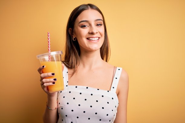 年轻美丽的黑发女人喝健康的橙汁在黄色的背景与快乐的脸站着，微笑着自信的微笑显示牙齿图片下载