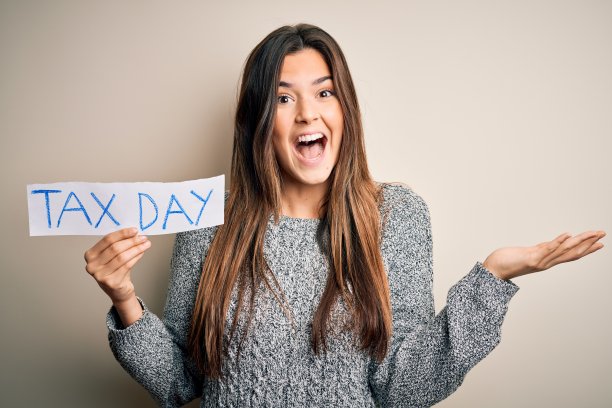 年轻漂亮的女孩拿着带有纳税日信息的纸张站在白色背景前，非常高兴和兴奋，以胜利的表情尖叫着庆祝胜利，露出灿烂的笑容并举起双手图片下载