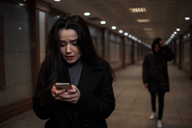 一个年轻漂亮的女人在晚上走过一个地下通道，后面跟着一个穿着深色衣服、戴着兜帽的男人。一位妇女试图拨打救援服务的电话图片下载