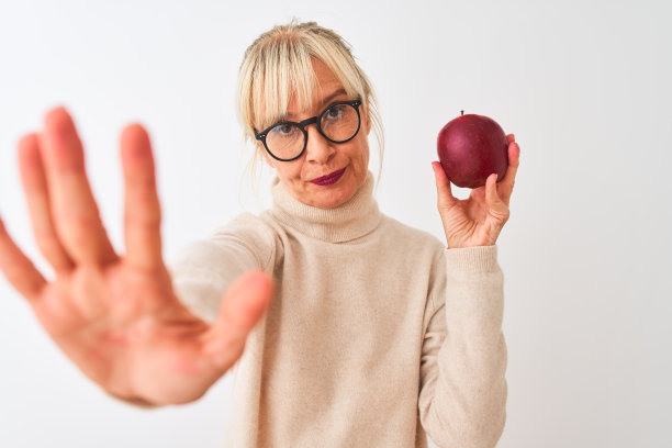 戴眼镜的中年妇女拿着苹果，在白色背景前做停止手势，表情严肃自信图片下载