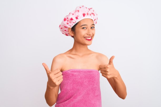 年轻的中国女子穿着浴巾和帽子沐浴在孤立的白色背景下，用手做积极的手势，竖起大拇指，微笑着为成功而高兴。赢家的姿态。图片下载
