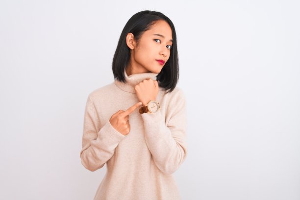 一名年轻的中国女子穿着高领毛衣站在孤立的白色背景下，匆忙地指着手表时间，不耐烦地看着镜头，神情轻松图片下载