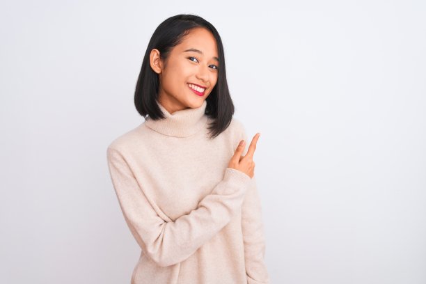 年轻的中国妇女穿着高领毛衣站在孤立的白色背景愉快地微笑着指着与手和手指向上的一边与快乐和自然的表情图片下载