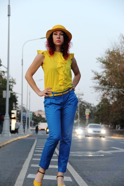 在路上，留着红色卷发，穿着蓝色经典长裤、黄色衬衫和帽子的女人。城市夜晚，远处有车灯图片下载