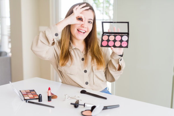 美丽的年轻女子使用化妆化妆，从调色与快乐的脸，微笑做ok手势与手在眼睛透过手指看图片下载