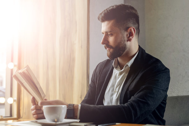 成年络腮胡男子在咖啡馆内，生活方式概念照片，专业人士在窗边看书喝咖啡图片下载